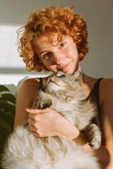 portrait red-haired curly young woman with pet gray fluffy cat in arms, lit by bright sun