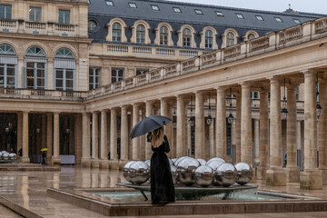 Obraz premium A blonde woman dressed in black with a black umbrella outside the ancient Palais-Royal in Paris, France