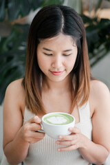 A young woman holding a cup of matcha latte, enjoying a peaceful moment in a cozy cafe with lush...