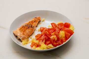 a white plate with grilled salmon, mashed potatoes and chopped tomato on a white veined marble table.