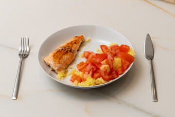 a white plate with grilled salmon, mashed potatoes and chopped tomato on a white veined marble table with a stainless stell fork and fish knife