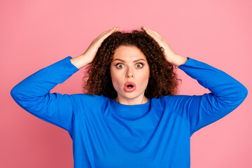 Surprised young woman with curly hairstyle in blue sweater against pink background showing shock or excitement