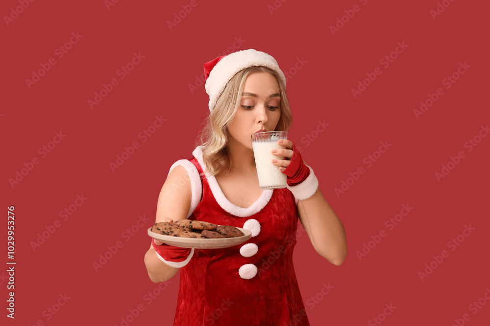 Wall mural Beautiful young woman dressed as Santa Claus drinking milk with tasty cookies on red background