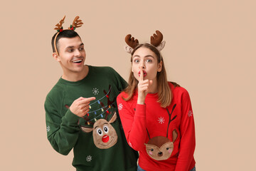 Beautiful young couple in reindeer horns showing silence gesture on beige background