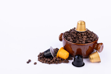 A brown bowl brimming with coffee beans, with a capsule on top, surrounded by loose beans and colorful coffee capsules, on a white background
