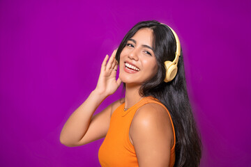 Fototapeta premium Side view of a smiling woman with yellow headphones, wearing an orange top, enjoying music and posing playfully against a purple background. 