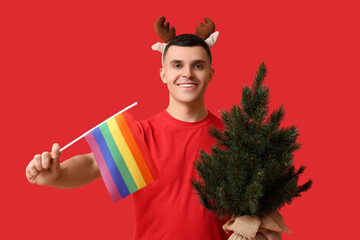 Young man in reindeer horns with LGBT flag and Christmas tree on red background