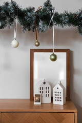 Ceramic house shaped lanterns, blank frame and Christmas decorations on wooden cabinet near white wall