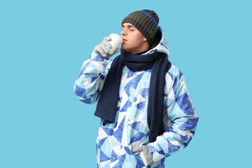 Young man in stylish puffer jacket and scarf with paper cup of coffee on blue background