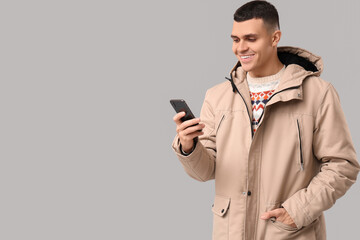 Young man in stylish puffer jacket with mobile phone on grey background