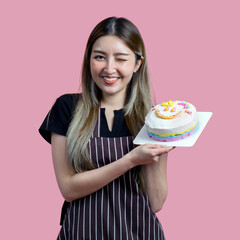 Young asian woman with an apron in celebratory or festive mood holding colorful cake with various sprinkles on the topping. Portrait on pink background with studio light. Isolated