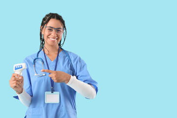 Young African-American female doctor with stethoscope pointing at thermometer on blue background