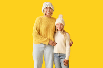 Beautiful woman and her cute daughter in stylish sweaters and hats on yellow background