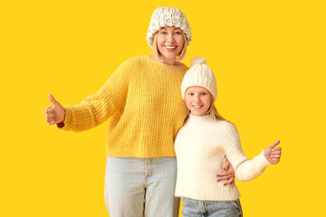 Beautiful woman and her cute daughter in stylish sweaters and hats showing thumb-up gesture on yellow background