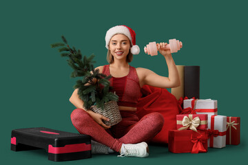 Sporty young woman with dumbbell, Christmas tree, gift boxes and aerobic stepper on green background