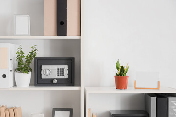 Modern safe, houseplants and office folders on shelving unit near white wall