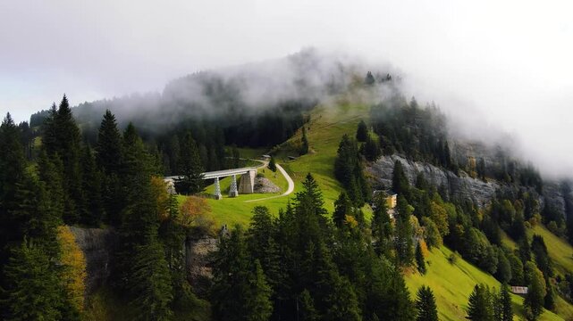 A cinematic drone shot of a misty alpine landscape with a winding path and railway bridge cutting through lush green hills and dense forests and fog-covered mountain peaks in Switzerland 