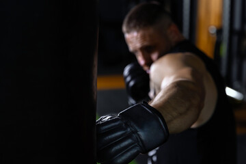 Focus on foreground closeup boxer with punching bag for training. A strong male fighter with boxing gloves trains on a punching bag in the gym.