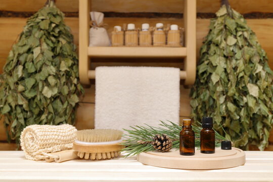 On a plank bench are a wooden circle tray with two bottles of healing essential oil next to a green pine branch and pine cone, set of bath supplies on the background of birch brooms and shelf. 