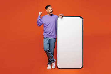 Full body young fun man wear purple sweatshirt casual clothes big huge blank screen mobile cell phone smartphone with area do winner gesture isolated on plain red orange background. Lifestyle concept.