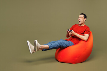 Full body young shocked fun middle eastern man wear red t-shirt casual clothes sit in bag chair hold in hand play pc game with joystick console isolated on plain green background. Lifestyle concept. © ViDi Studio