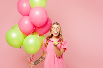 Young fun excited woman wear shirt celebrating birthday holiday and hold bunch of colorful air balloons look at small cake with candle isolated on plain pastel light pink background Lifestyle concept