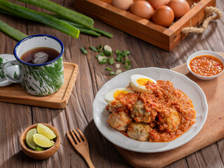 A plate of siomay, an Indonesian dish, topped with a spicy peanut sauce and boiled egg halves, served alongside lime wedges and a dipping sauce. A cup of tea and fresh ingredients are placed