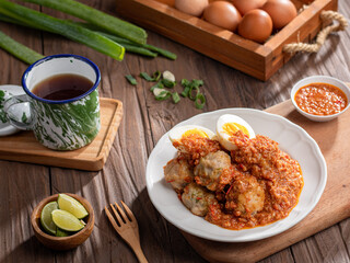 A plate of siomay, an Indonesian dish, topped with a spicy peanut sauce and boiled egg halves, served alongside lime wedges and a dipping sauce. A cup of tea and fresh ingredients are placed