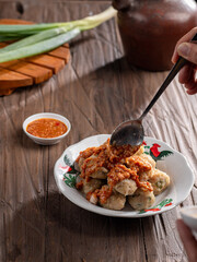 A plate of Indonesian siomay topped with peanut sauce and sweet soy sauce, served with dipping sauce. Nearby are a traditional teapot, cup, and fresh green onions, all set on a wooden surface.