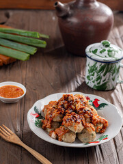 A plate of Indonesian siomay topped with peanut sauce and sweet soy sauce, served with dipping sauce. Nearby are a traditional teapot, cup, and fresh green onions, all set on a wooden surface.