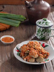 A plate of Indonesian siomay topped with peanut sauce and sweet soy sauce, served with dipping sauce. Nearby are a traditional teapot, cup, and fresh green onions, all set on a wooden surface.