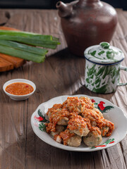 A plate of Indonesian siomay topped with peanut sauce and sweet soy sauce, served with dipping sauce. Nearby are a traditional teapot, cup, and fresh green onions, all set on a wooden surface.