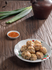 A plate of steamed Indonesian siomay served with a side of dipping sauce. Fresh green onions and a traditional teapot are placed nearby on a rustic wooden table, creating a simple yet inviting scene.