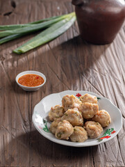 A plate of steamed Indonesian siomay served with a side of dipping sauce. Fresh green onions and a traditional teapot are placed nearby on a rustic wooden table, creating a simple yet inviting scene.