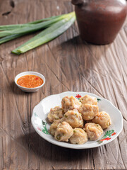 A plate of steamed Indonesian siomay served with a side of dipping sauce. Fresh green onions and a traditional teapot are placed nearby on a rustic wooden table, creating a simple yet inviting scene.