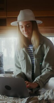 A young woman in a hat smiles while opening a laptop and starting typing while sitting on a bed in a van