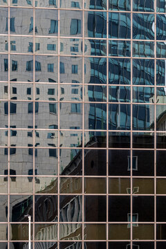 The glass facade of a building reflects an abstract, distorted view of the surrounding architecture, creating a dynamic representation of modern urban life.