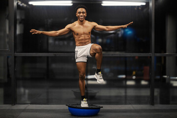 Smiling muscular shirtless strong diverse sportsman balancing on one foot at gym on bosu ball.