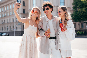 Man and two young beautiful smiling hipster female. Carefree women posing outdoors. Positive models holding and drinking fresh cocktail drink in glass bottle. In the street. Take selfie photos