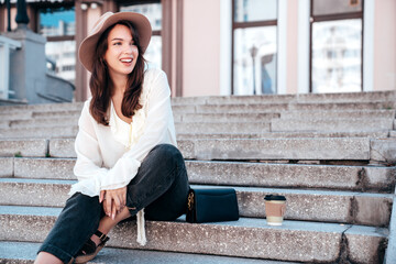 Young beautiful smiling hipster woman in trendy summer white blouse and jeans. Carefree woman posing in the street in sunny day. Positive model outdoors. Cheerful and happy, sits at stairs