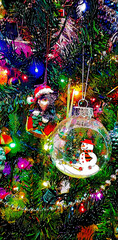 A festive Christmas tree adorned with colorful decorations. Prominent features include a glass ornament shaped like a snow globe containing a small snowman and a whimsical ornament of a cat.