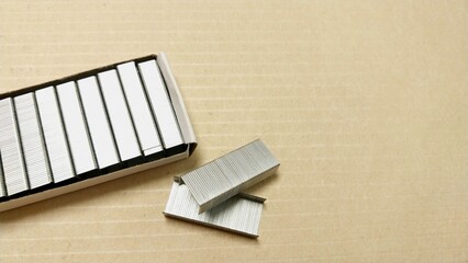 a box of staples on a beige, textured surface. The box is open, revealing several rows of neatly stacked staples inside. A few staples are also lying outside the box on the surface.