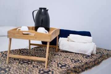 Bedroom setup with a wooden tray, black jug, cups, and rolled towels on a patterned bedspread.