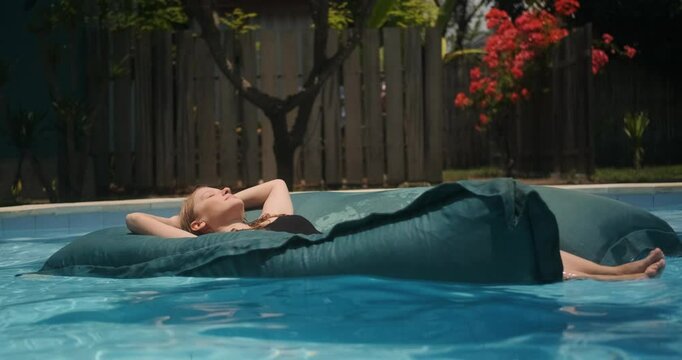 Young woman floating on a mattress in a pool with her eyes closed and enjoying her vacation on a tropical island. Relaxation and wellness treatments at all inclusive hotel. Spa treatments and swimming