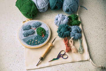Beautiful embroidered landscape in a frame placed on piece of fabric together with woolen skeins, punch needle and scissors