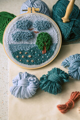 Punch needle embroidery in a hoop with blue and green balls of wool and a handle of punch needle in...