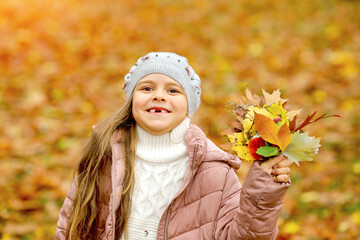 Cute Caucasian girl, smile without front teeth, in a knitted gray biret with a bouquet of autumn leaves. Autumn park background blur. Childhood concept