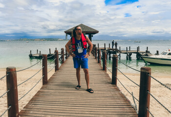 A tourist enjoys a day by the beach in Malaysia, posing confidently on a wooden pier surrounded by calm waters and boats