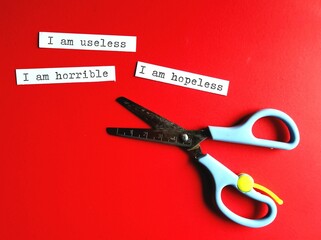 Scissors on red background with text paper written I AM HOPELESS I AM USELESS I AM HORRIBLE,...