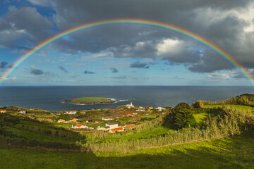 Fototapeta premium Breathtaking view of the rainbow over the Atlantic coastline with a small lighthouse and island on the background in Sao Jorge Azores Portugal